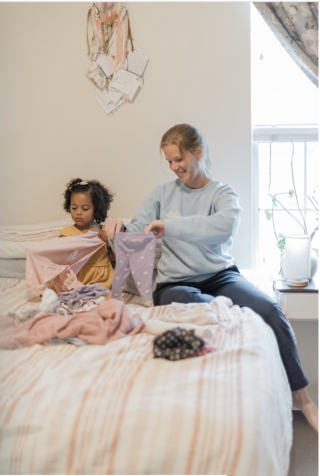 Kristin folding baby clothes on a bed beside a young child
